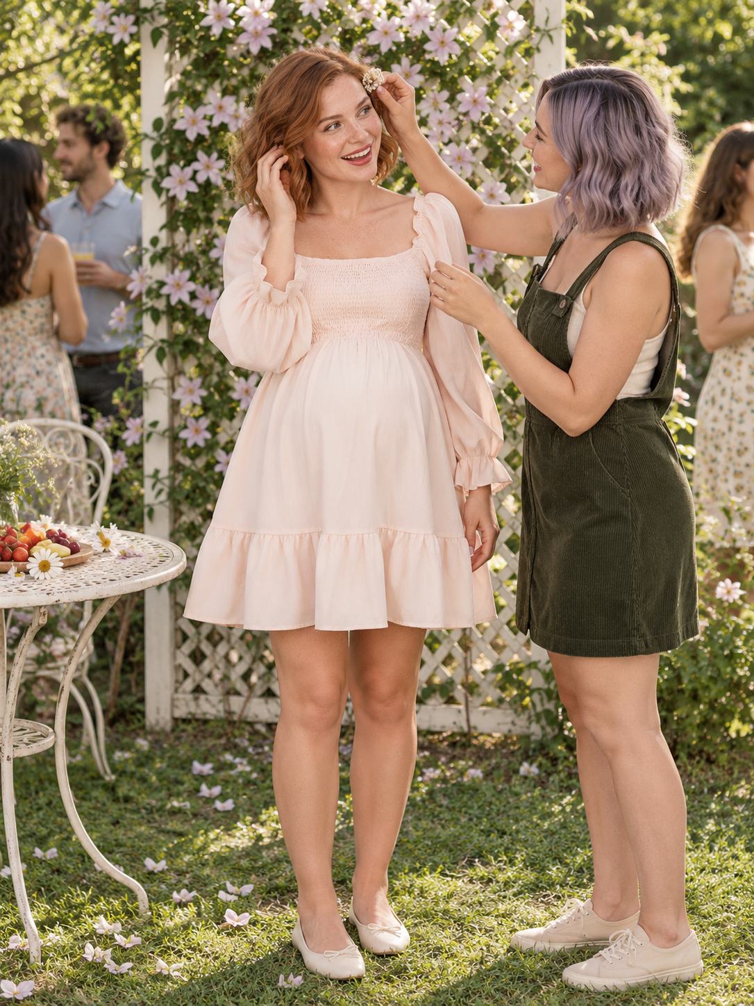 Penelope Pale Blush maternity dress — standing on the lawn as a friend adjusts a flower in her hair