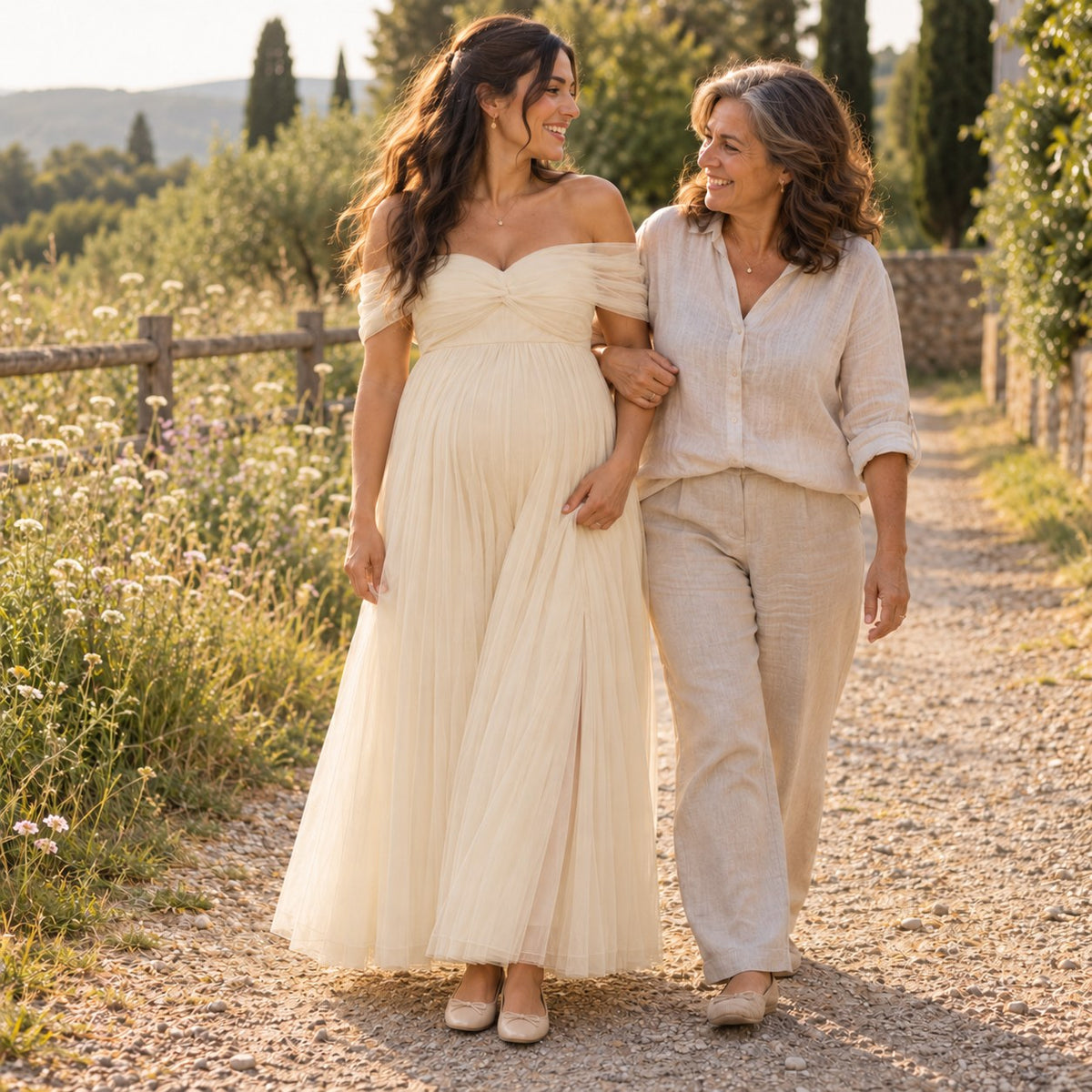 Mom-to-be in a champagne tulle maternity gown walking arm in arm with her mother beside a wildflower meadow