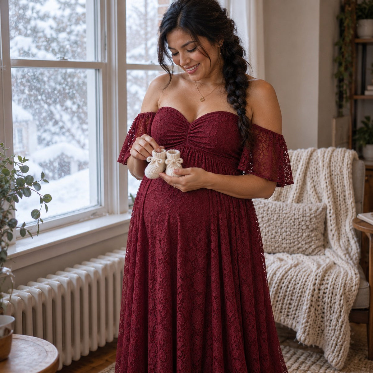 Mom-to-be in a wine red lace dress holding tiny knitted baby booties by a snowy window
